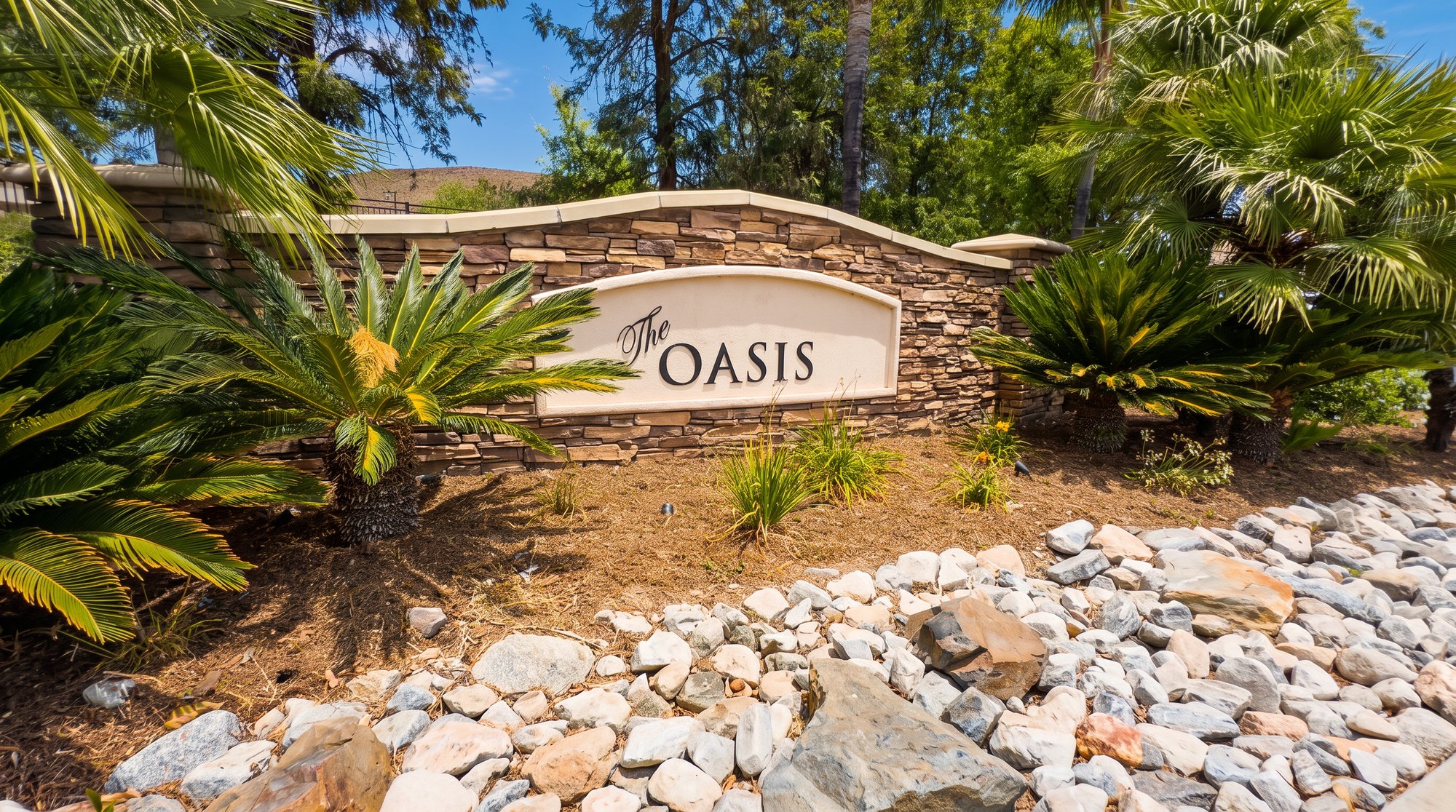 The stone entry monument at The Oasis, a gated 55+ community in Menifee, framed by palms and boulders